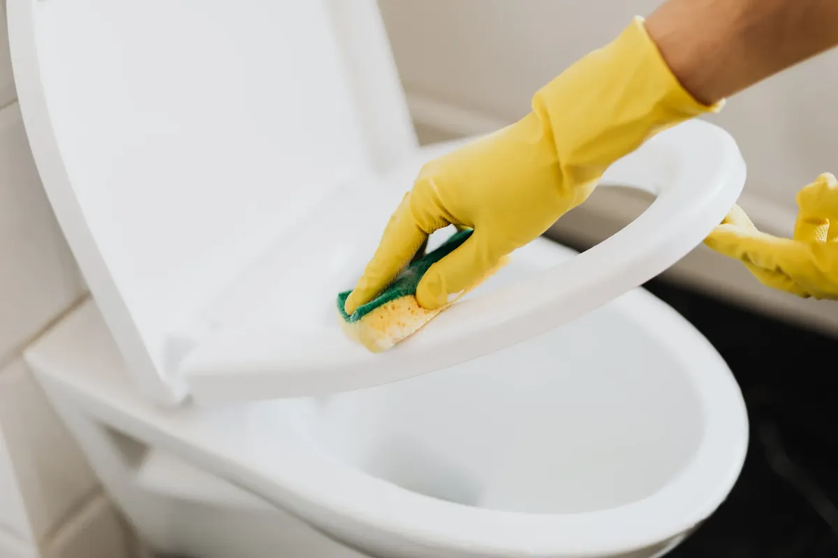 上面から、顔のない人物が黄色のゴム清掃手袋を着けたまま、pongeを使ってプラスチックのトイレットシートを適切に清掃している