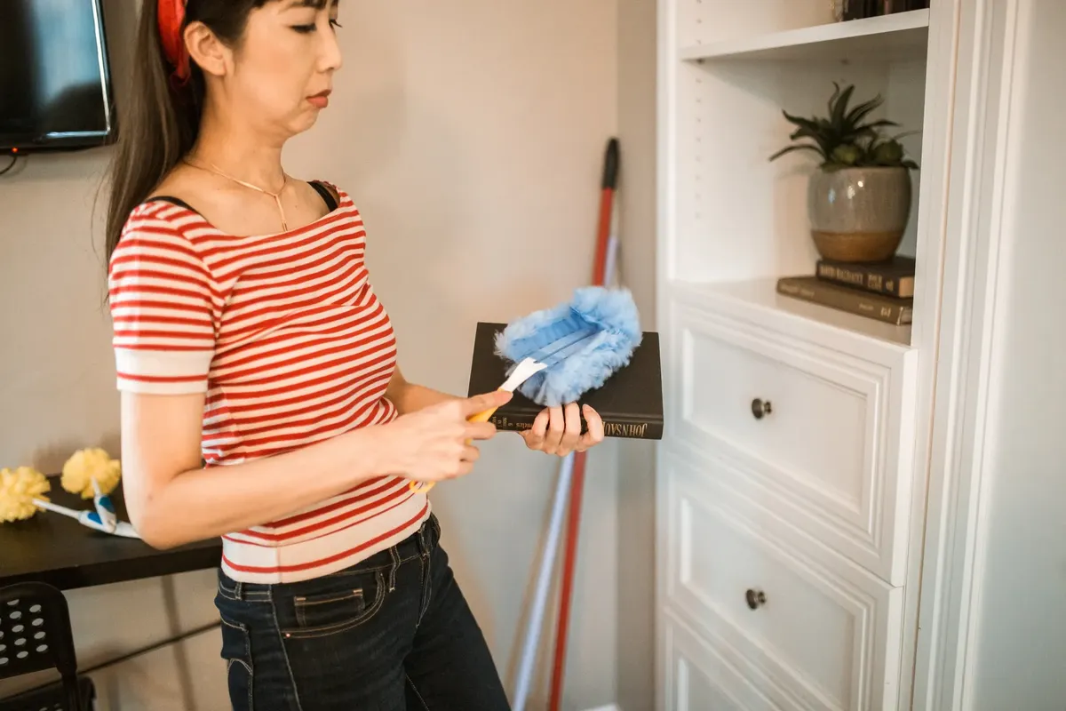 A woman dusting a bookshelf with a blue duster as part of house chores. Indoors setting.