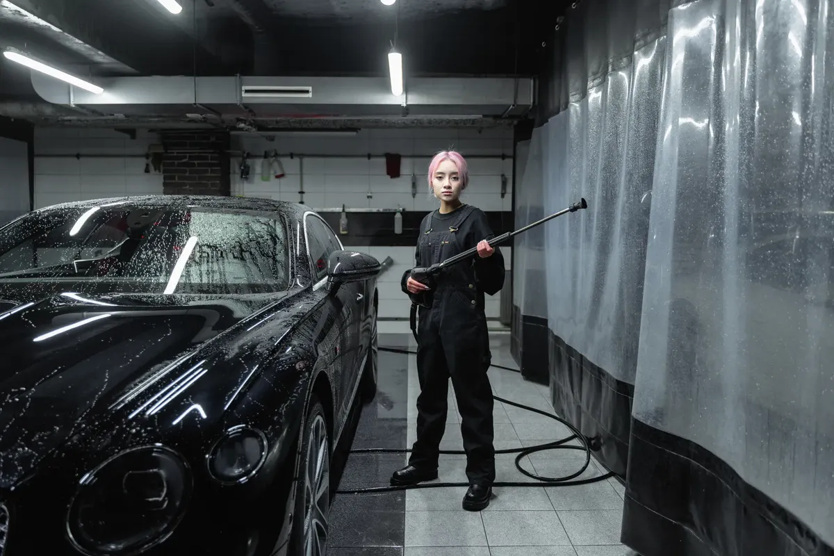 A woman using a pressure washer to clean a black sports car at an indoor car wash facility.