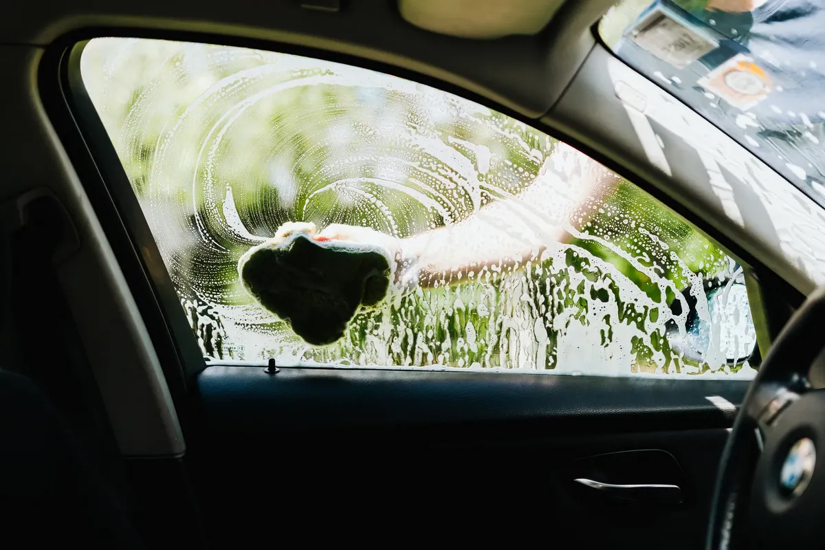 Driver's side car window being washed with a soapy sponge outdoors, capturing the cleaning process.